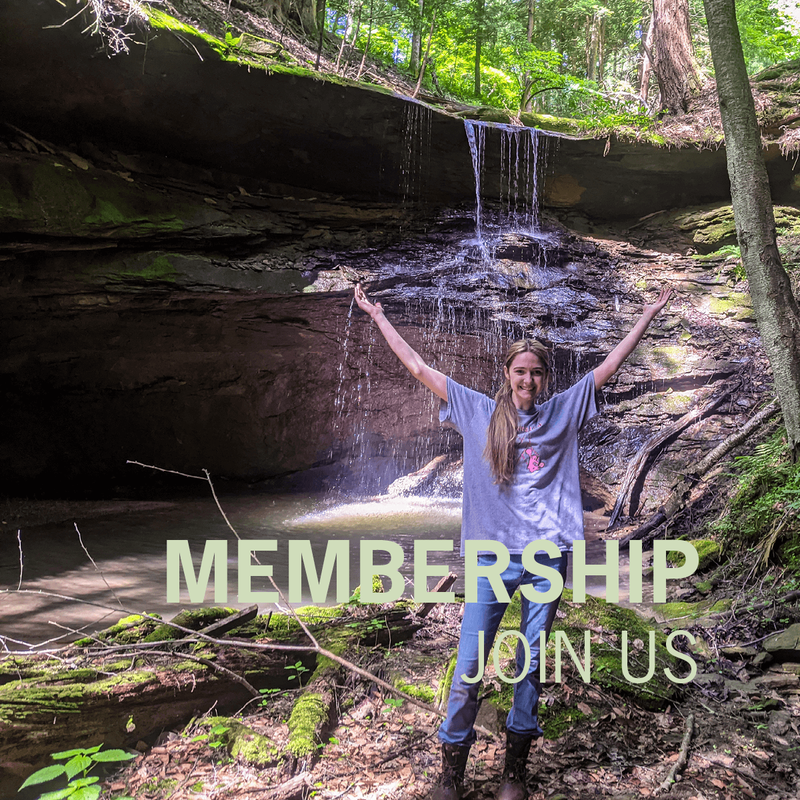 A woman with long blond hair in a low side ponytail wearing a gray t-shirt, jeans, and rain boots is holding her arms outstretched above her head while standing in front of a waterfall. The light green text “Membership, Join Us” is overlaid in the bottom right corner.