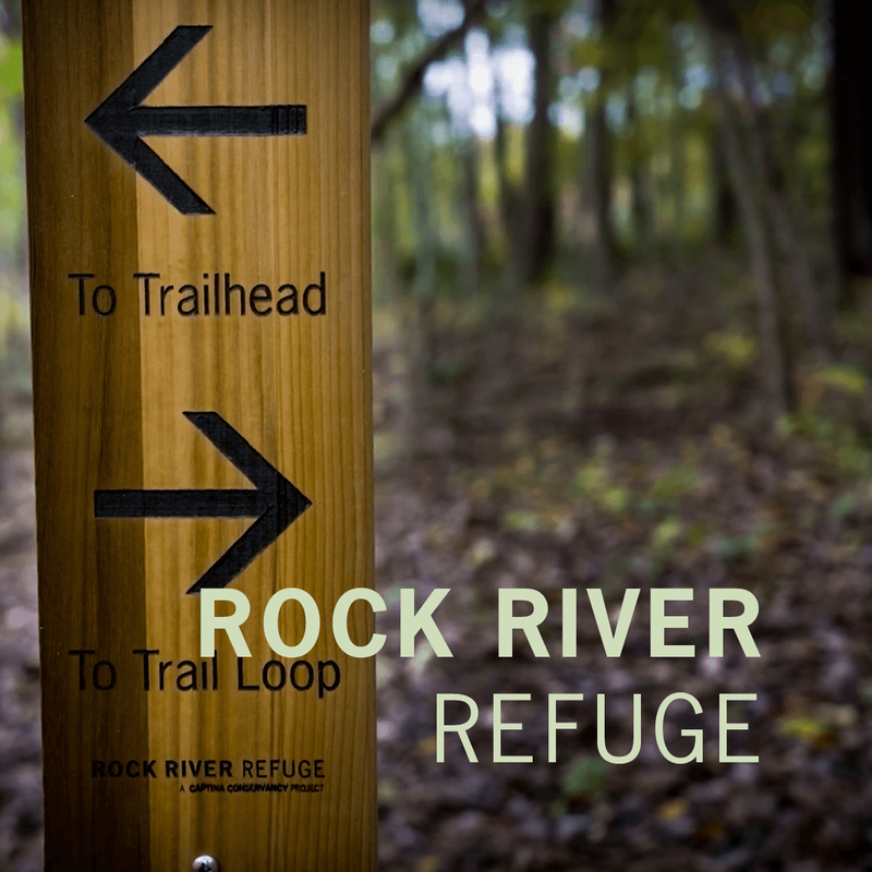A long rectangular wooden sign with two arrows pointing in opposite directions is staked in a forest. The light green text “Rock River Refuge” is overlaid in the bottom right corner.
