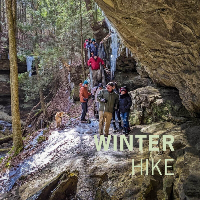 People are walking under a rock overhang on a cold, snowy day. The light green text “Winter Hike” is overlaid in the bottom right corner.
