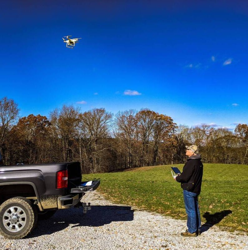 A man wearing a ball cap, a long-sleeved black jacket, and blue jeans is holding a remote control unit while he looks upward at a small white drone that is hovering in the cloudless, blue sky. The back half of a black pickup truck is to the left of the man.